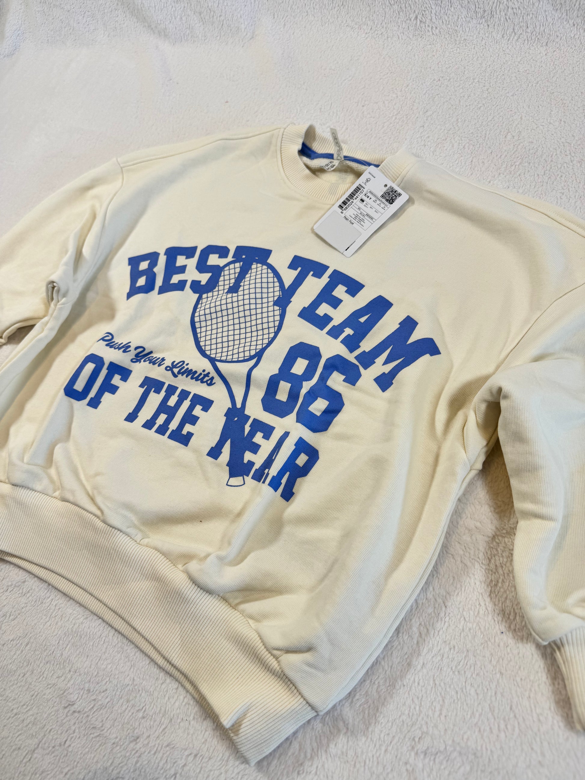 Cream-colored sweatshirt with blue tennis-themed graphic and text, featuring the phrase "110/116 Schöner Pullover Jungs" on the tag, laid on a soft white surface.