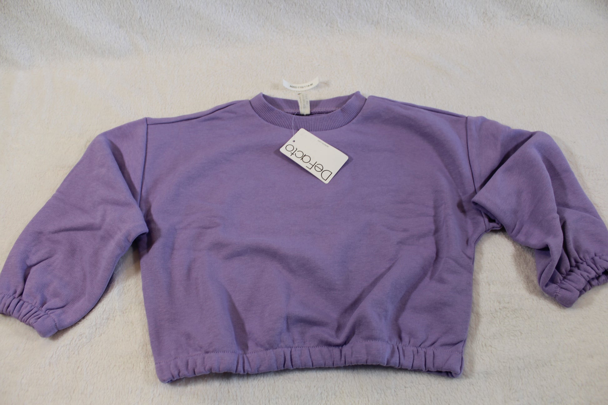 110/116 schöner Uni Pullover in soft purple fabric laid flat on a light surface with a tag attached, featuring long sleeves and elastic cuffs.
