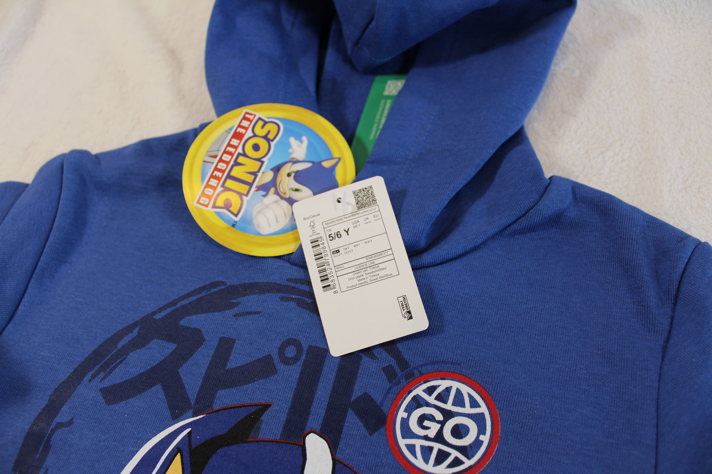 Close-up of a blue 110/116 Pullover Sonic hoodie with a graphic design and attached retail tags on a light fabric background.